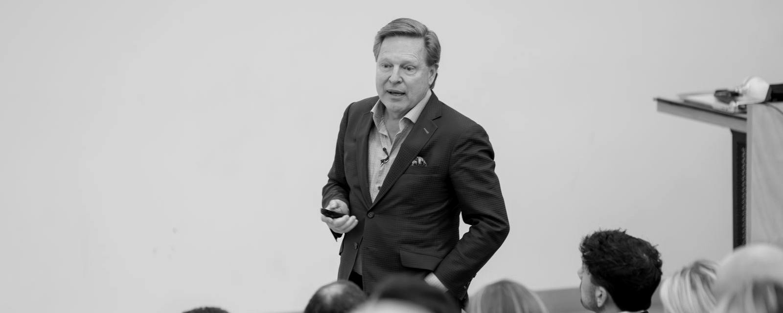 Verne Harnish, em preto e branco, de pé diante de uma plateia de executivos, segurando um controle de slides
