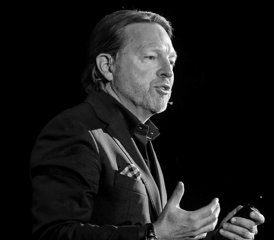 Verne Harnish de perfil, falando ao microfone de lapela num palco escuro, gesticulando com a mão